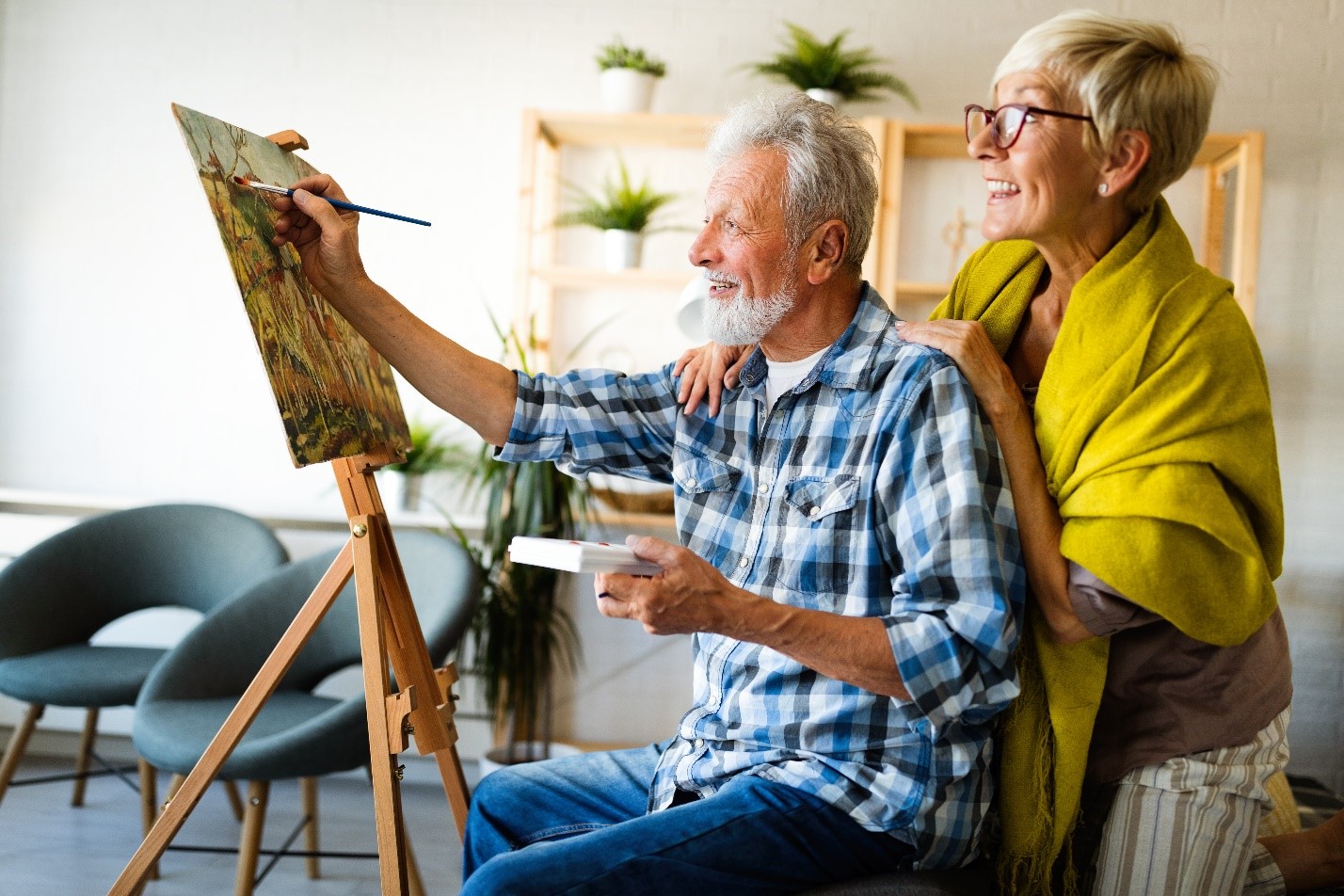55 and older man painting on an easel while a woman cheers him on behind him